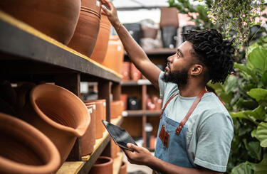 Man assessing different clay pots, dressed in a sage green short sleeved t-shirt and a blue apron with brown leather straps 