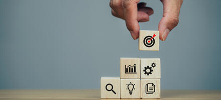 Small building blocks GettyImages-1370817574 Hand using small building blocks each with an image of a lightbulb, magnifying glass, documents, cogs, chart and bullseye